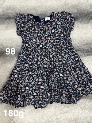 Blommig klänning i marinblått - Supersöt klänning med volanger och blommigt mönster i ljusa färger på marinblå botten. Klänningen har korta ärmar och rund hals. Perfekt för sommarens alla tillfällen!🌸 Fint använt skick. Storlek 98