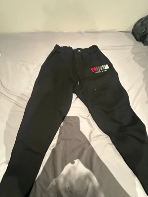 Svarta mjukisbyxor med broderi - Svarta mjukisbyxor från Trapstar med broderad logga i rött, grått och vitt på vänster lår. Byxorna har snörning i midjan och dragkedjor vid bensluten för extra stil. Tillverkade i mjukt bomullsmaterial som är skönt att ha på sig.