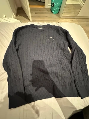 Mörkblå stickad tröja Bondelid XL - Snygg mörkblå stickad tröja från Bondelid i storlek XL. Tröjan har ett klassiskt kabelstickat mönster, rund hals och broderad logga med röd sköld på bröstet. Perfekt för dig som gillar stilrena och tidlösa plagg med lite preppy känsla.