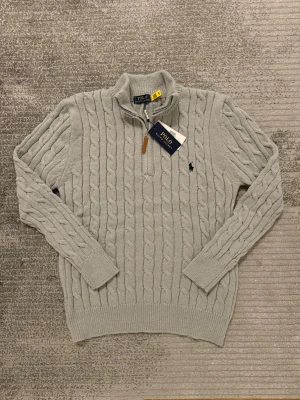 Ljusgrå stickad tröja Polo Ralph Lauren - Säljer en ljusgrå kabelstickad tröja från Polo Ralph Lauren med half zip och hög krage. Tröjan har lång ärm, ribbade muddar och den klassiska broderade loggan på bröstet. Perfekt för dig som gillar stilren och tidlös design.