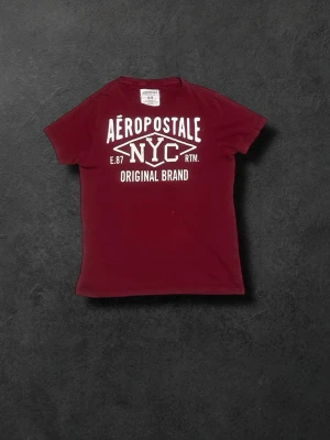 Vinröd Aeropostale NYC t-shirt - Snygg vinröd t-shirt från Aeropostale med stort NYC-tryck och texten 'Original Brand' framtill. Klassisk rund hals och korta ärmar. Tillverkad i 100% bomull för en skön och avslappnad känsla. Perfekt för dig som gillar streetstyle och enkel design.