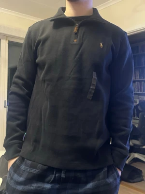 Svart half zip tröja från Polo Ralph Lauren - Säljer en svart långärmad tröja från Polo Ralph Lauren med klassisk broderad logga på bröstet. Tröjan har en snygg half zip-dragkedja med läderdetalj och hög krage. Tillverkad i mjuk bomull, perfekt för lager på lager under höst och vinter.