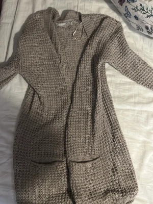 Grå stickad kofta med fickor - Mysig grå stickad kofta i längre modell med två praktiska fickor framtill. Grovstickad struktur och ribbade muddar vid ärmslut. Perfekt att slänga över en t-shirt under kyliga dagar.