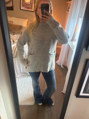 Grå stickad tröja med fickor - Mysig grå stickad tröja med två fickor framtill och ribbade muddar. Tröjan har en loose passform, lång ärm och liten slits i sidorna. 