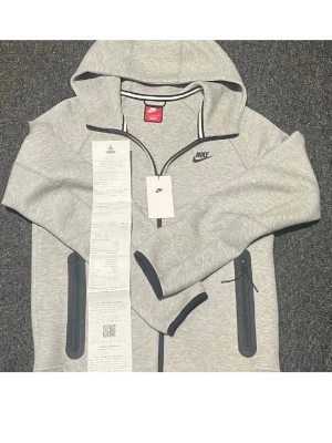 Nike tech  - Säljer en stilren grå hoodie från Nike med dragkedja och huva. Jackan har svarta detaljer och två praktiska fickor med dragkedjor. Perfekt för en sportig look eller avslappnade dagar. Orginal kvitto och orginal prislapp ingår. Såklart äkta skriv vid minsta lilla funderinga. Den är helt ny den har bara legat i min garderob den är bara testad en gång annars helt ny. Nypris 1250