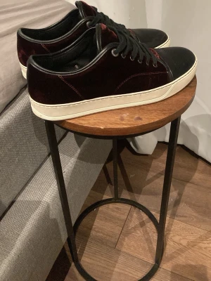 Lanvin sneakers i sammet och skinn - Säljer ett par Lanvin sneakers med vinröd sammet på sidorna och svart skinn på tån. Skorna har vita gummisulor och svarta skosnören. Klassisk låg modell med rund tå och exklusiv känsla. Perfekta för dig som vill ha något unikt och stilrent.