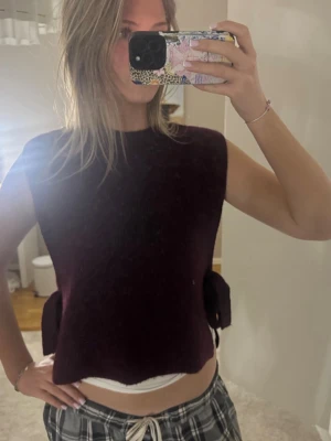 Vinröd stickad ärmlös tröja med knyt - Säljer en vinröd stickad ärmlös tröja med rund hals och snygga knytband i sidorna. Tröjan har en loose passform och är perfekt att styla över en t-shirt eller skjorta. Supermjuk och passar grymt till höst och vinter.