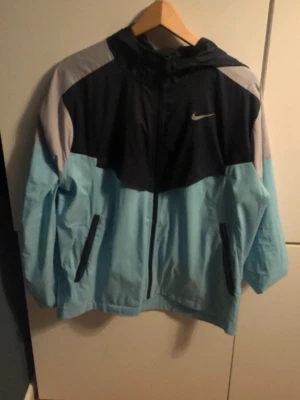 Nike Miller jacka  - Snygg vindjacka från Nike i ljusblått, svart och grått. Jackan har huva, dragkedja framtill och två fickor med svarta detaljer. Perfekt för dig som vill ha en sportig och stilren look.