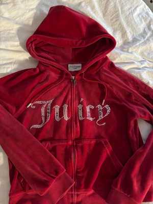 Röd Juicy Couture velourväst - Juicy Couture i röd velour med huva och dragkedja. Stora glittriga stenar bildar texten 'Juicy' på bröstet. Klassisk juicy-stil med fickor framtill och mjukt material som känns lyxigt.