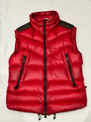 Röd dunväst från Moncler Grenoble - Säljer en äkta röd dunväst från Moncler Grenoble med hög krage, svarta detaljer på axlarna och två snedställda fickor med dragkedja. Västen har quiltad design och justerbar nederkant med dragsko. Perfekt för lager på lager under kyliga dagar. 