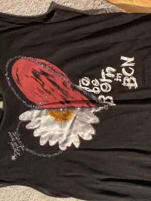 Svart ärmlös topp med blomtryck Desigual - Unik svart ärmlös topp från Desigual med stort tryck av en röd hjärtformad detalj och en vit prästkrage på framsidan, dekorerad med små paljetter. Texten 'to be born in BCN' och 'why?' finns tryckt på plagget. Baksidan har en liten prästkrage och knapp i nacken.