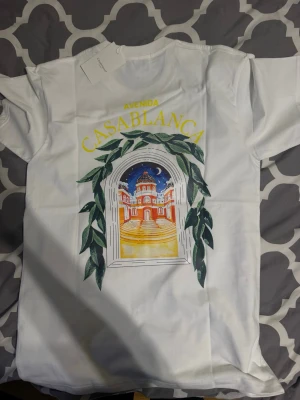  Casablanca t-shirt  - Vit t-shirt från Casablanca med färgglatt tryck framtill som visar en byggnad i solnedgång och gröna blad runt ett fönster. Texten 'Avenida Casablanca' i gult ovanför motivet. Klassisk passform och rund hals. Perfekt för dig som gillar statement-plagg. Helt ny storlek Small säljs för 900kr men priset går att diskutera 