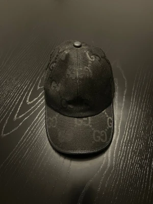 Svart Gucci keps  - Säljer en svart keps från Gucci med GG-mönster över hela kepsen. Tillverkad i 90% merinoull och har justerbar rem bak. Snygg och stilren accessoar med diskret logga och exklusiv känsla.