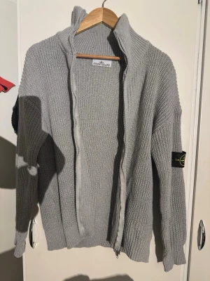 Grå stickad kofta från Stone Island - Säljer en grå stickad kofta från Stone Island med dragkedja framtill och klassisk patch på ärmen. Koftan har hög krage och ribbade muddar. Perfekt för lager på lager under höst och vinter. Snygg och enkel att matcha till det mesta.