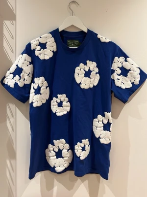  Denim Tears T-shirt  - Säljer en blå t-shirt från Denim Tears med stora vita blommiga mönster över hela tröjan. T-shirten har rund hals och är kortärmad. Materialet är mjuk bomull och passformen är normal. Perfekt för dig som vill sticka ut med en unik look. Perfekt 