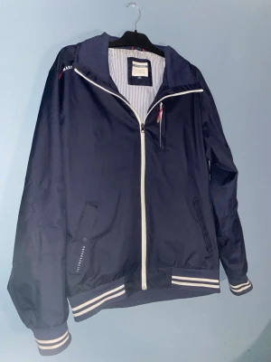 Marinblå vindjacka från Race Marine XL - Snygg marinblå vindjacka från Race Marine i storlek XL. Jackan har vita dragkedjor, randiga muddar och krage samt två fickor framtill. Insidan är fodrad med vitt och blått randigt tyg. Perfekt för dig som gillar sportig och clean stil.