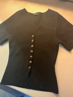 Svart kofta med v-ringning från Gina Tricot - Säljer en svart kofta från Gina Tricot med v-ringad hals och korta ärmar. Tröjan har snygga knappar framtill och är i ett mjukt material som känns skönt mot huden. Perfekt att styla till jeans eller kjol för en clean look.