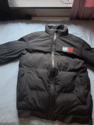 Svart pufferjacka Tommy Hilfiger S - Svart pufferjacka från Tommy Hilfiger med hög krage och dragkedja framtill. Jackan har två stora fickor med dragkedja och logga på bröstet samt stor logotyp på ryggen. Perfekt för kalla dagar och riktigt snygg streetstil.