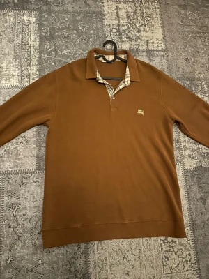 Brun pikétröja från Burberry - Säljer en brun långärmad pikétröja från Burberry med klassisk rutig krage och broderad logga på bröstet. Tröjan har en snygg knappslå och är perfekt för dig som gillar stilrena och tidlösa plagg.