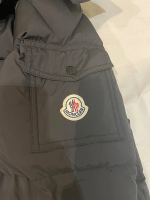 Svart Moncler dunjacka - Säljer en svart Moncler dunjacka med klassisk logga på ärmen och coolt serietryck på insidan. Jackan har dragkedja, tryckknappar och en ficka på ärmen. Perfekt för kalla dagar och riktigt snygg streetstil.
