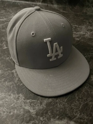 Grå LA-keps från New Era - Säljer en stilren grå keps från New Era med LA-logga broderad framtill i vitt. Klassisk modell med böjd skärm och ventilerande hål upptill. Perfekt accessoar för en sportig och avslappnad look.