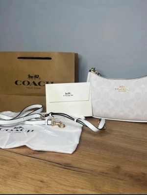 Vit axelväska från Coach - Säljer en stilren vit axelväska från Coach med diskret mönster och guldfärgade detaljer. Väskan har dragkedja upptill, avtagbar axelrem och Coach-logga i guld framtill. Tillverkad i skinn och har en klassisk, lätt rundad form. Köpte till sambon men hon hade tydligen beställt en likadan. 