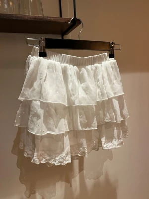 Vit volangkjol med spetsdetaljer - Supersöt vit kortkjol med flera lager volanger och spetskant längst ner. Kjolen har resår i midjan och är gjord i ett lätt och luftigt tyg. Helt ok skick! Se bilder. Passar xs-s. 