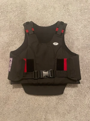 Svart ridväst  - Säljer en svart skyddsväst från Biltema i storlek C XL för barn/ungdom. Västen har röda detaljer, justerbara axelband och spänne framtill. Märkt med BETA 2009 Level 3 för ridning. Perfekt för dig som vill ha extra skydd vid ridning. Aldrig använt den så den är som helt ny!