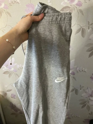 Grå mjukisbyxor från Nike - Säljer ett par grå mjukisbyxor från Nike med klassisk logga broderad i vitt på benet. Byxorna har resår i midjan med snörning och är gjorda i mjukt bomullsmaterial. Perfekta för chill eller träning.