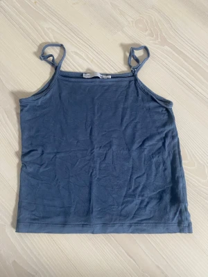 Blåt linne från 157 - En enkel blå linnetopp från 157 med tunna axelband och rak passform. Toppen är gjord i mjuk bomull och har en stilren look utan mönster eller detaljer. Perfekt för varma dagar eller som lager under en skjorta. Storlek 140.