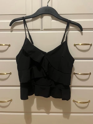 Svart volangtop - Säljer en svart linnetopp med tunna justerbara axelband och snygga volangdetaljer framtill. Toppen har en croppad passform och är perfekt för dig som gillar en enkel men ändå trendig look.