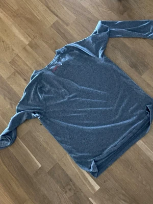 Grå långärmad träningstopp Under Armour - Säljer en grå långärmad träningstopp från Under Armour. Toppen har en stor logga och text framtill i rött och vitt. Tillverkad i ett mjukt och stretchigt syntetmaterial som andas, perfekt för träning eller sport.