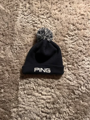 Svart PING mössa med tofs - Snygg svart stickad mössa från PING med vit broderad logga framtill och en svartvit tofs på toppen. Perfekt accessoar för kalla dagar och passar till de flesta outfits. Klassisk design med uppvikt kant. Också fett grish golf mössa !!!