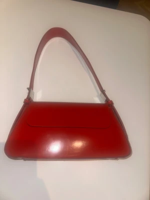 Röd axelväska i skinn från Zara - Snygg röd axelväska från Zara i glansigt skinn. Väskan har en stilren, avlång form med magnetlås och justerbar axelrem. Invändigt finns ett fack och svarta textildetaljer. Perfekt för dig som vill sticka ut med en färgklick till din outfit.