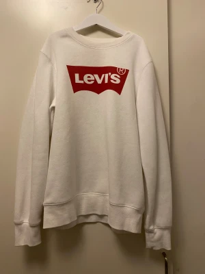 Vit sweatshirt från Levi's med röd logga - Vit sweatshirt från Levi's med stor röd logga på bröstet. Tröjan har rund hals, långa ärmar och ribbade muddar vid ärmslut och nederkant. Mjuk insida och klassisk passform, perfekt för en chill och stilren look.