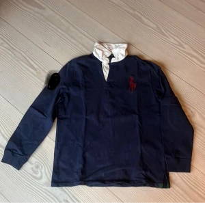 Mörkblå pikétröja Polo Ralph Lauren - Snygg mörkblå långärmad pikétröja från Polo Ralph Lauren med vit krage och gröna detaljer. Röd broderad logga på bröstet och nummer 3 på ryggen. Tillverkad i mjuk bomull, perfekt för dig som gillar klassisk sportig stil. Använd 1 gång och är  en speciell tröja/kofta.