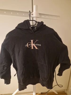 Svart hoodie Calvin Klein Jeans - Svart hoodie från Calvin Klein Jeans med stor CK-logga i ljusrosa och vitt framtill. Klassisk känguruficka, dragsko i huvan och ribbade muddar. Mjuk bomullskänsla och avslappnad passform, perfekt för en chill look.
