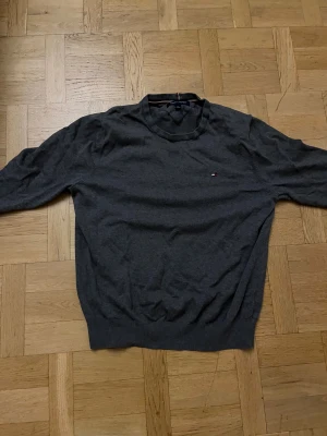 Mörkgrå tröja Tommy Hilfiger M - Snygg mörkgrå tröja från Tommy Hilfiger i storlek M. Klassisk design med rund hals och diskret logga på bröstet. Tröjan har långa ärmar och ribbade muddar vid ärmslut och nederkant. Perfekt för dig som gillar stilrena och enkla plagg.