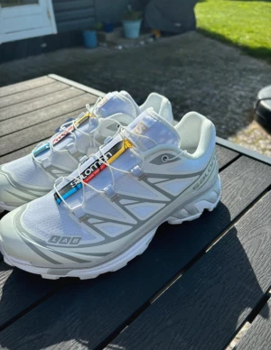  Salomon XT-6 sneakers - Säljer ett par riktigt snygga Salomon XT-6 sneakers i vitt med grå detaljer. Skorna har mesh-överdel, coola färgdetaljer på plösen och snabbsnörning. Perfekta för dig som gillar streetwear och outdoor-vibe. Sulan är rejäl och ger bra grepp. Märkta med LAB-logga på sidan.