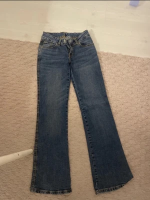 Blå bootcut jeans med snörning - Snygga blå jeans med bootcut passform och cool snörning i midjan. Klassisk femficksmodell med slitningar och kontrastsömmar. Perfekta för dig som gillar en retro vibe och vill ha lite extra attityd i din outfit.