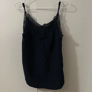 Svart linnetopp med spets från ONLY - Säljer en svart linnetopp från ONLY med tunna axelband och vacker spetsdetalj längs urringningen. Toppen har en slät, mjuk yta och en feminin, enkel design som passar perfekt till jeans eller kjol.
