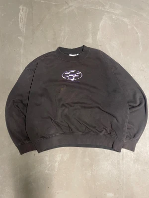 Grå sweatshirt med lila tryck - Cool svart sweatshirt med ett lila och vitt grafiskt tryck på bröstet där det står 'PERFORMANCE 12.05.80'. Tröjan har ribbade muddar vid ärmslut och midja samt en klassisk rund halsringning. Materialet känns som bomull och passformen är oversized.