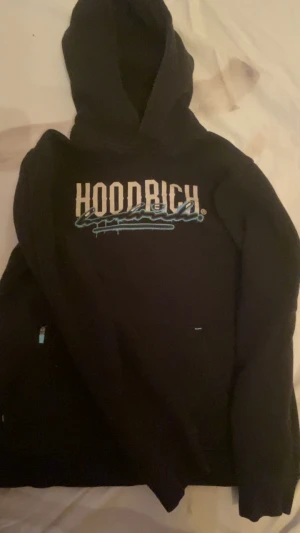 Svart hoodie från Hoodrich - Säljer en svart hoodie från Hoodrich med stor vit och blå logga på bröstet. Hoodien har huva och en klassisk känguruficka framtill. Materialet känns mjukt och bekvämt, perfekt för chill dagar. Snygg streetwear-stil med coolt tryck.
