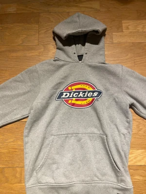 Grå Dickies hoodie med logga - Snygg grå hoodie från Dickies i storlek small. Stor färgglad Dickies-logga på bröstet i rött, gult och blått. Tröjan har huva, magficka och ribbade muddar. Perfekt för chill dagar.
