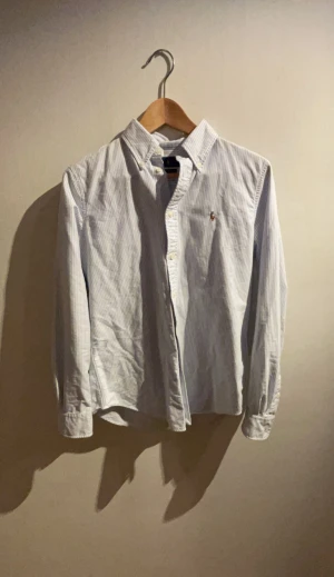 Randig skjorta från Ralph Lauren M - Säljer en klassisk vit och ljusblå randig skjorta från Ralph Lauren i custom fit. Skjortan har button-down krage, broderad logga på bröstet och långa ärmar med knappar vid ärmslut. Perfekt för en clean och stilren look.