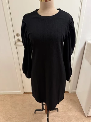 Svart stickad klänning från JDY - Svart stickad klänning från JDY i storlek medium. Modellen är kort med rund hals och långa puffärmar som ger en snygg siluett. Materialet är stickat och har en diskret struktur. Perfekt för dig som gillar enkel men trendig stil.