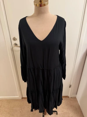 Svart volangklänning Jacqueline de Yong - Svart klänning från Jacqueline de Yong i storlek 40. Modellen har v-ringning, långa ärmar och flera volanglager som ger en flowy känsla. Klänningen är kort och har en avslappnad passform, perfekt för dig som gillar en enkel men snygg stil.