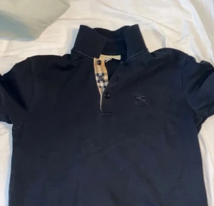 Svart pikétröja Burberry XS - Snygg svart pikétröja från Burberry i storlek L. Klassisk krage och korta ärmar, med den ikoniska rutiga Burberry-detaljen vid knäppningen och två svarta knappar. Tröjan är gjord i mjuk bomull och har en diskret Burberry-logga broderad på bröstet.