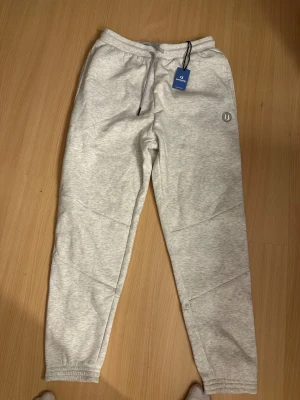 Ljusgrå mjukisbyxor från Unisport - Ljusgråa mjukisbyxor från Unisport i storlek M. Byxorna har snörning i midjan, ribbad mudd och en bakficka med dragkedja. Materialet är mjukt och skönt och är endast testade. 