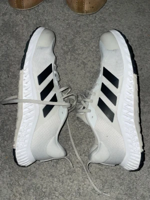 Vita Adidas träningssneakers - Säljer ett par vita Adidas träningssneakers med svarta ränder på sidorna och grov svart yttersula. Skorna har snörning och är tillverkade i syntetmaterial och mesh för bra ventilation. Snygg sportig look med platt sula och rund tå, perfekta för gymmet eller löpning.